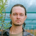 A man with long brown hair, blue eyes, and a short beard stands outdoors in front of a green leafy plant and a blue-tinted background, wearing a plaid shirt over a beige t-shirt.