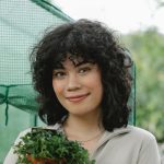 A person with curly dark hair smiles while holding a small potted plant. They are standing outdoors near a green netted structure with greenery visible in the background.
