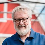 An older man with white hair, a full white beard, and glasses smiles while wearing a blue collared shirt. A red building with white trim and a glass roof is in the blurred background.