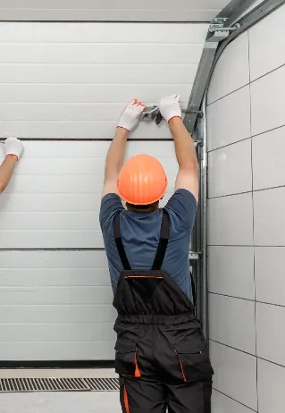 A worker in overalls and an orange hard hat adjusts a mechanism on a white garage door from inside a garage, using tools and wearing white gloves.
