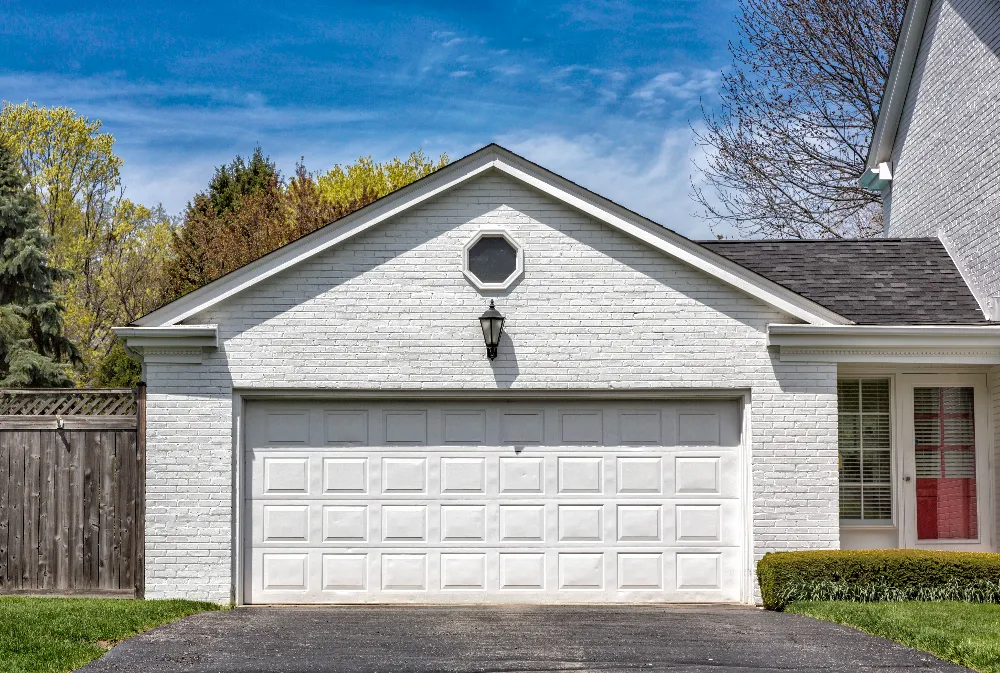 A white brick house with a double garage door, a black lantern light above the door, and a small round window. Trees and a wooden fence are in the background under a blue sky.