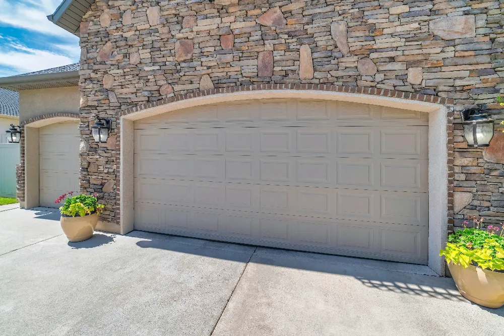 Beige double garage door on a stone exterior house, with potted pink and red flowers on either side and wall-mounted lantern-style lights above each flower pot. Concrete driveway in front.