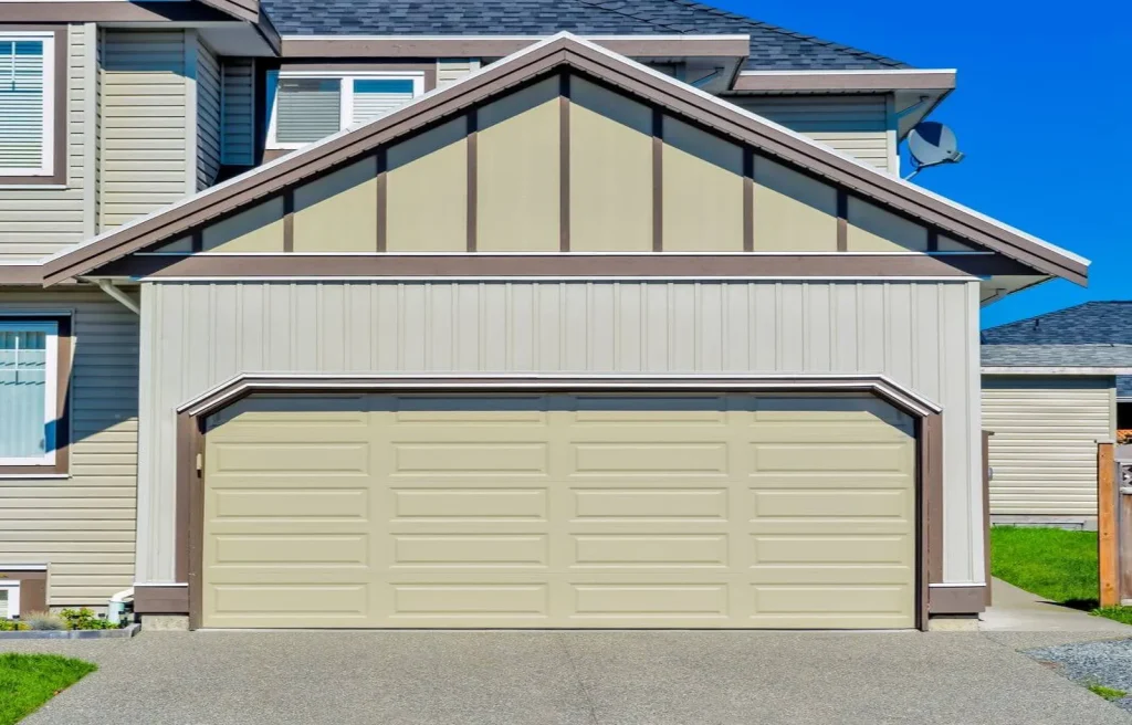A beige two-car garage with a closed door, attached to a modern house. The exterior features light siding with brown trim, and the driveway is concrete under a clear blue sky.