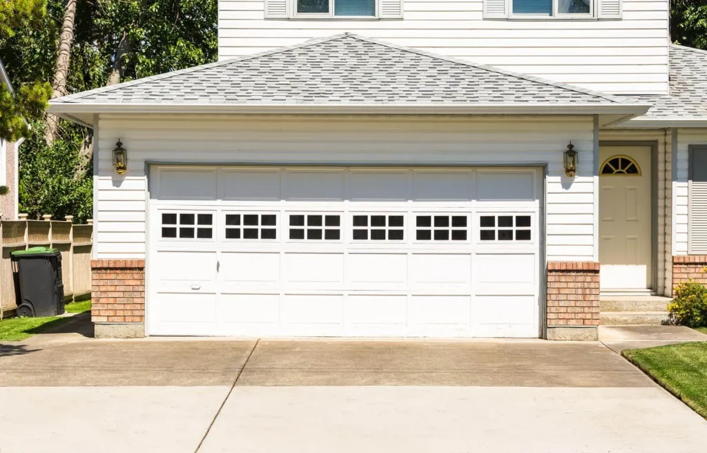 A clean white garage door with small windows at the top, attached to a house with white siding, brick accents, and a concrete driveway. There are lamps on each side and a side door to the right.