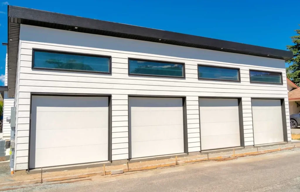 A modern white building with three garage doors and three narrow rectangular windows above them, set against a clear blue sky.