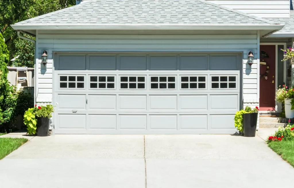A light gray double garage door with rectangular windows along the top row, flanked by black planters with green plants, set against a white house and a concrete driveway.