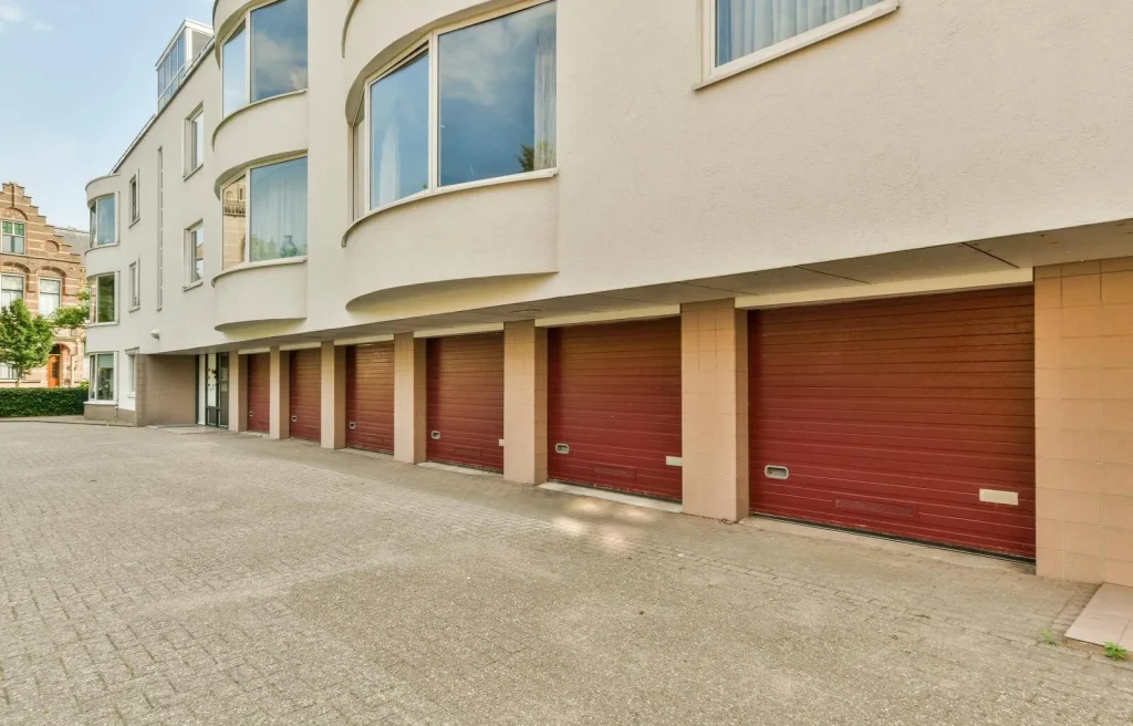 A row of five closed red garage doors beneath a modern cream-colored building with large curved windows and a paved driveway in front.