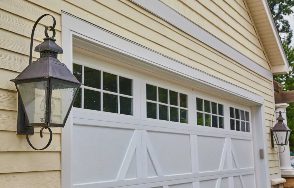 A close-up view of a white garage door with windows, attached to a yellow-sided house. Two black metal lantern-style light fixtures are mounted on either side of the garage door.