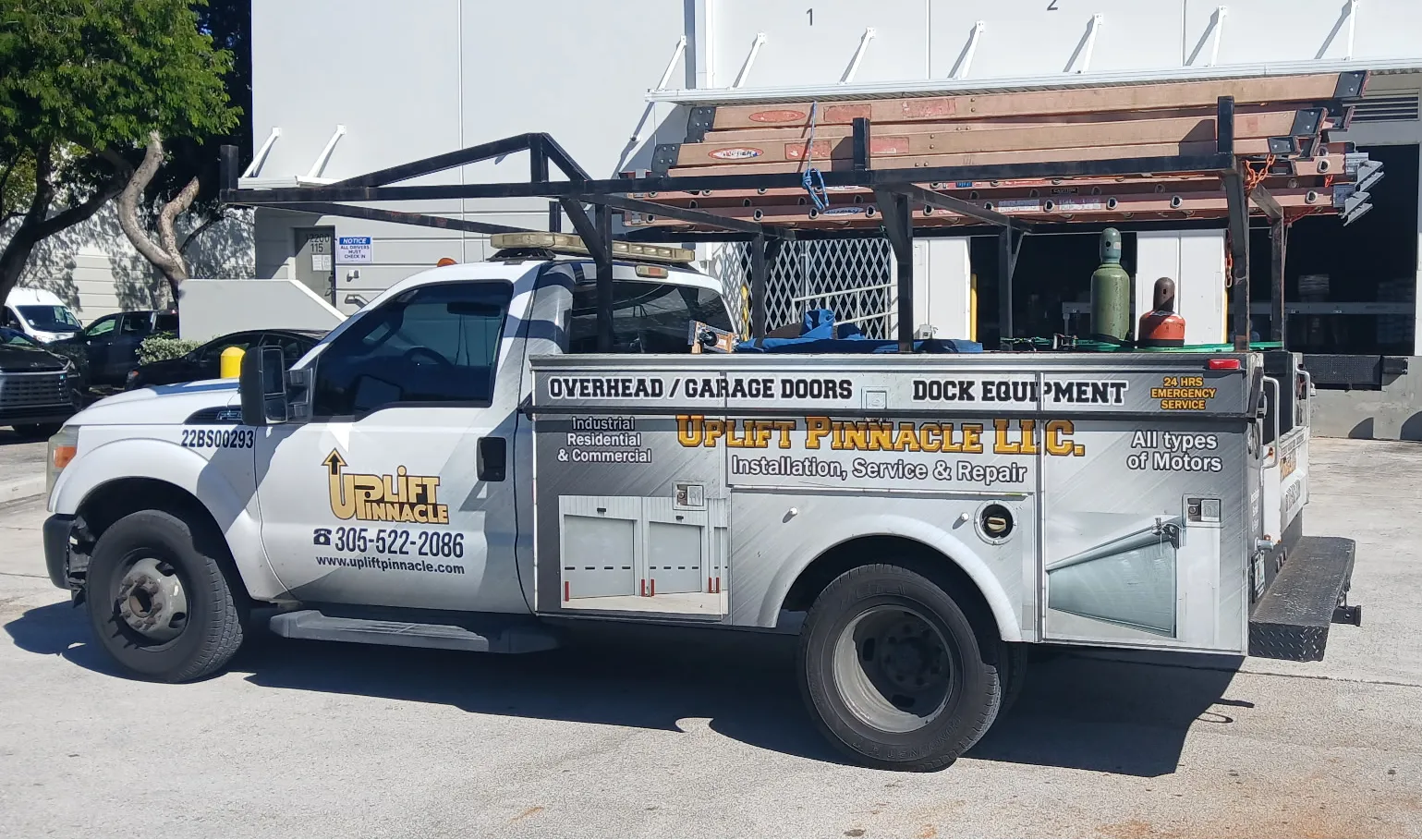 A white service truck with racks and equipment on top is parked outside a building. The truck advertises "Uplift Pinnacle LLC" for overhead/garage doors and dock equipment installation, service, and repair.
